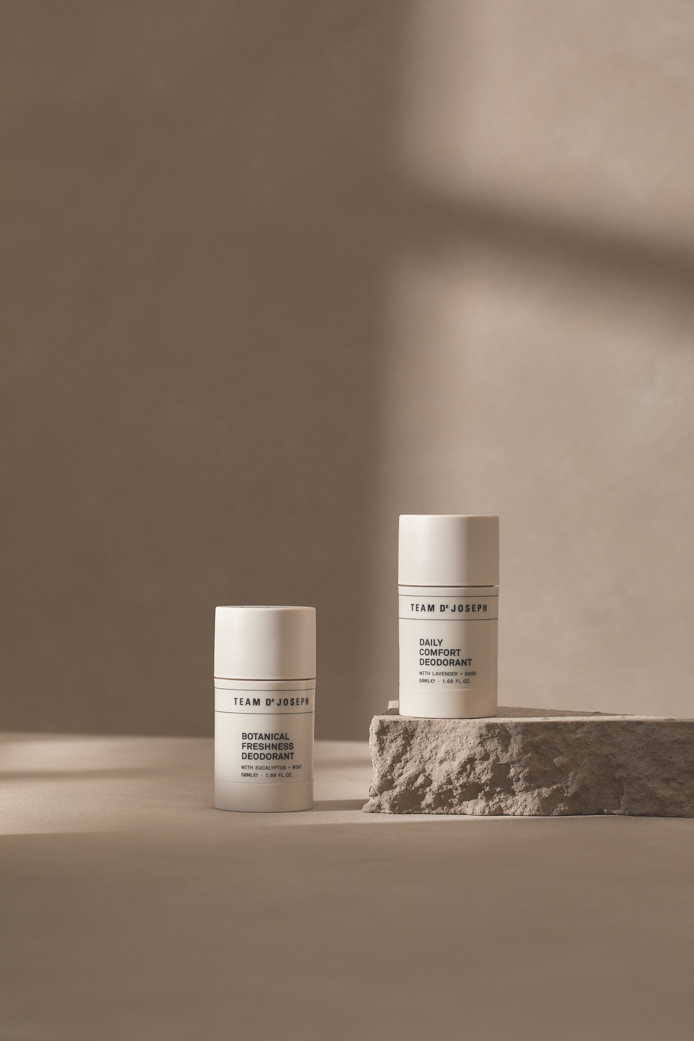 Two white deodorant bottles on a neutral surface, one on a stone block, against a muted beige background with a subtle shadow.