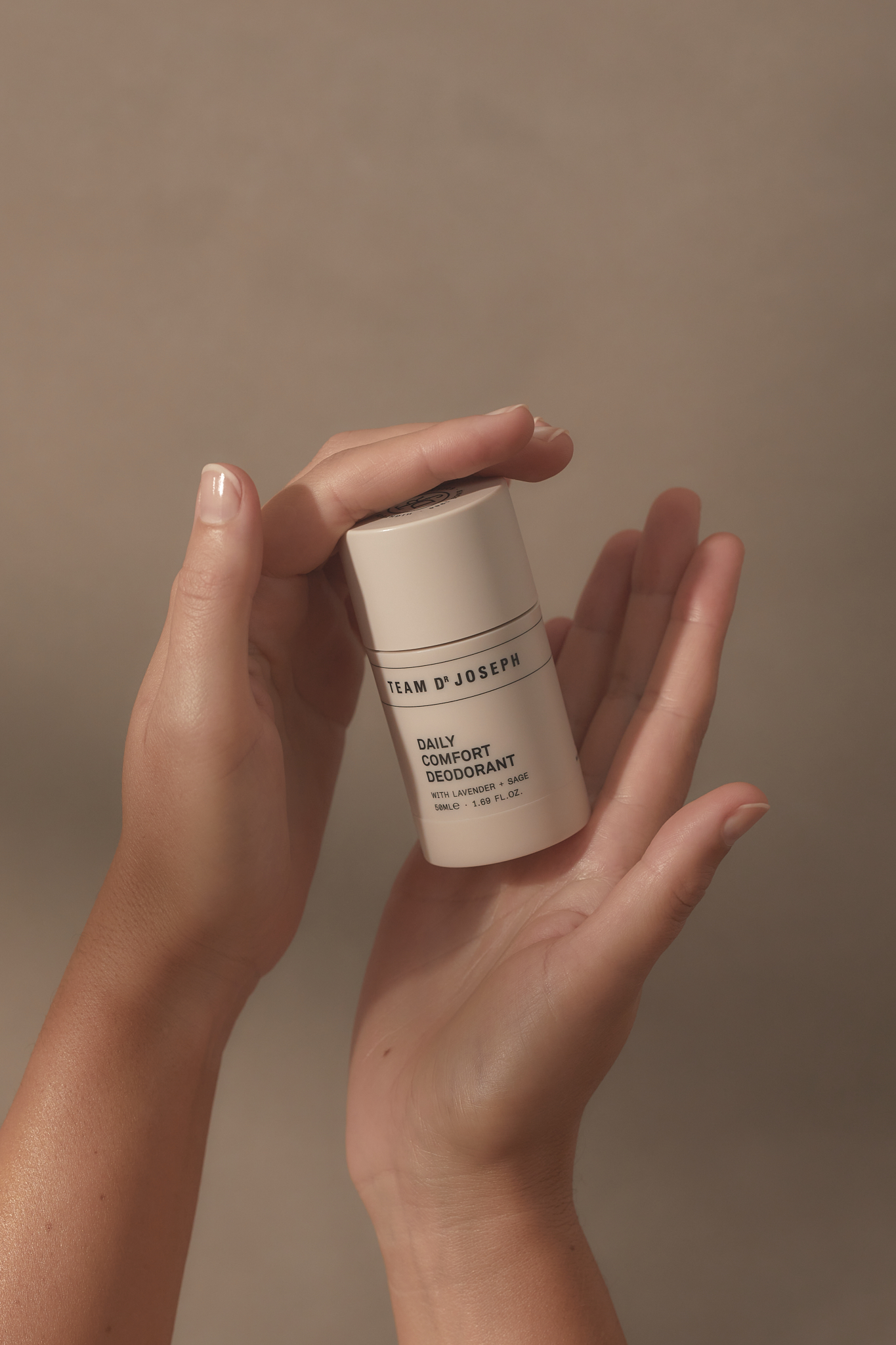 Hands hold white tube of Team D’Josephs Daily Comfort Deodorant product against neutral backdrop in a beauty and skincare product shot.