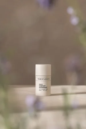 Team Dr Joseph Daily Comfort Deodorant stick standing on a neutral surface framed by soft purple flowers.