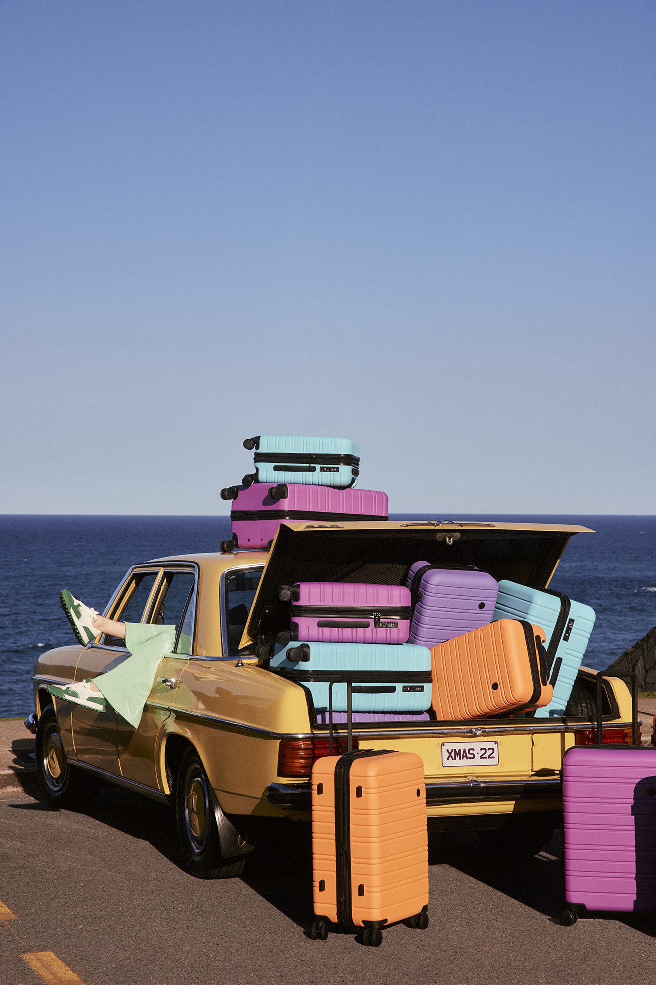 Yellow vintage car trunk full of pink and blue luggage on oceanside road under blue sky. Suitcases stacked on top of car.