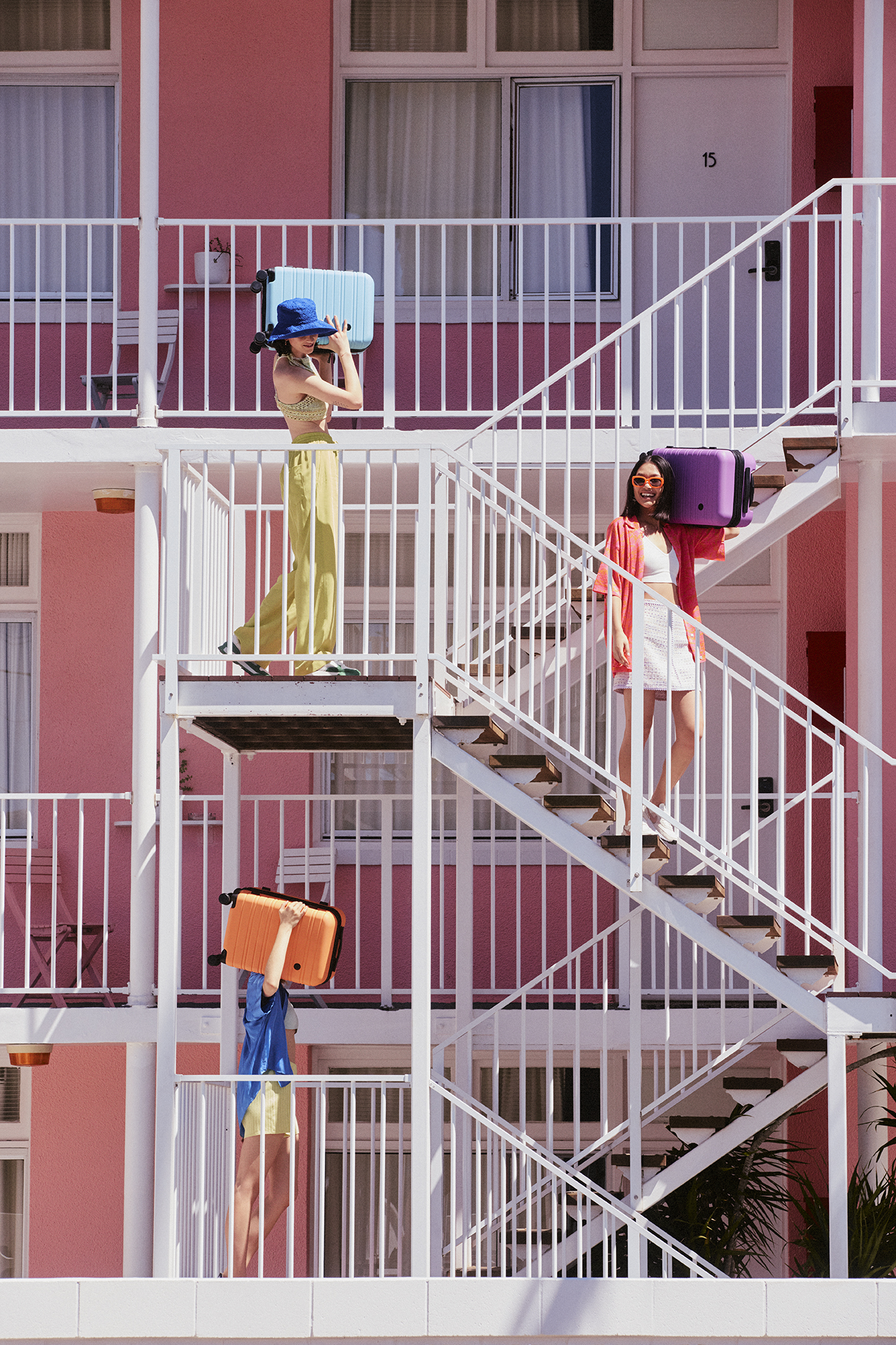 Three women on white outdoor stairs of a pink hotel, each carrying colorful luggage. Travel and fashion concept.