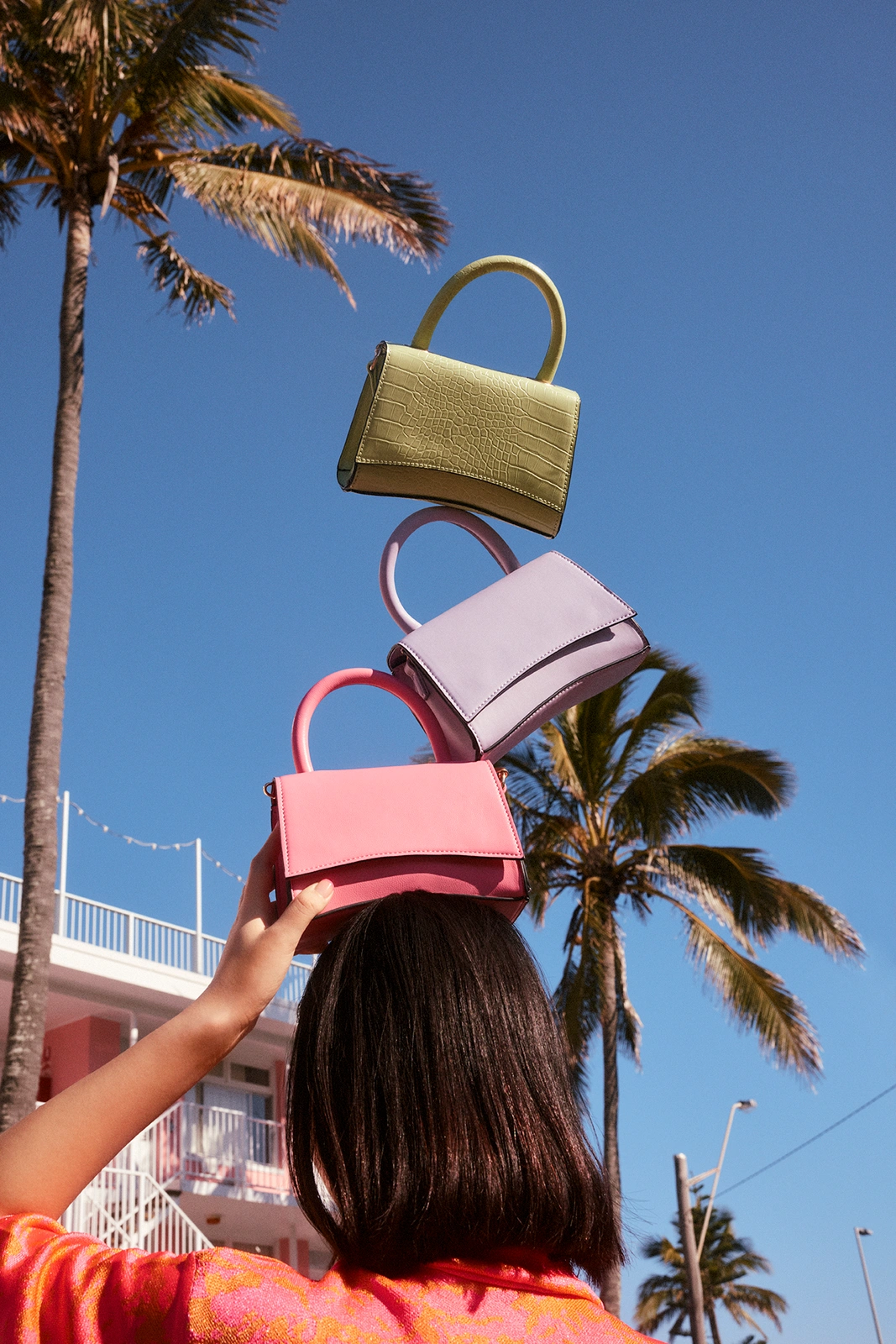 A person balancing a stack of pastel handbags on their head against a blue sky with palm trees for Strand Bags Summer