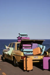 Yellow vintage car packed with colorful suitcases by the ocean under a clear blue sky, with a person's legs resting out the window