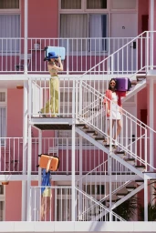 Three models carrying suitcases on their heads walk on exterior white metal staircases against a vibrant pink building.