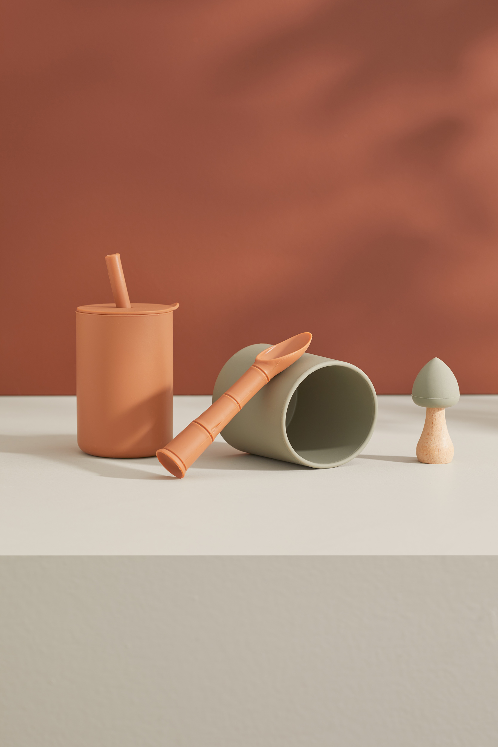 Still life shot of cups, a spoon, and a mushroom-shaped toy on a white surface, set against a two-tone background with a shadow effect.