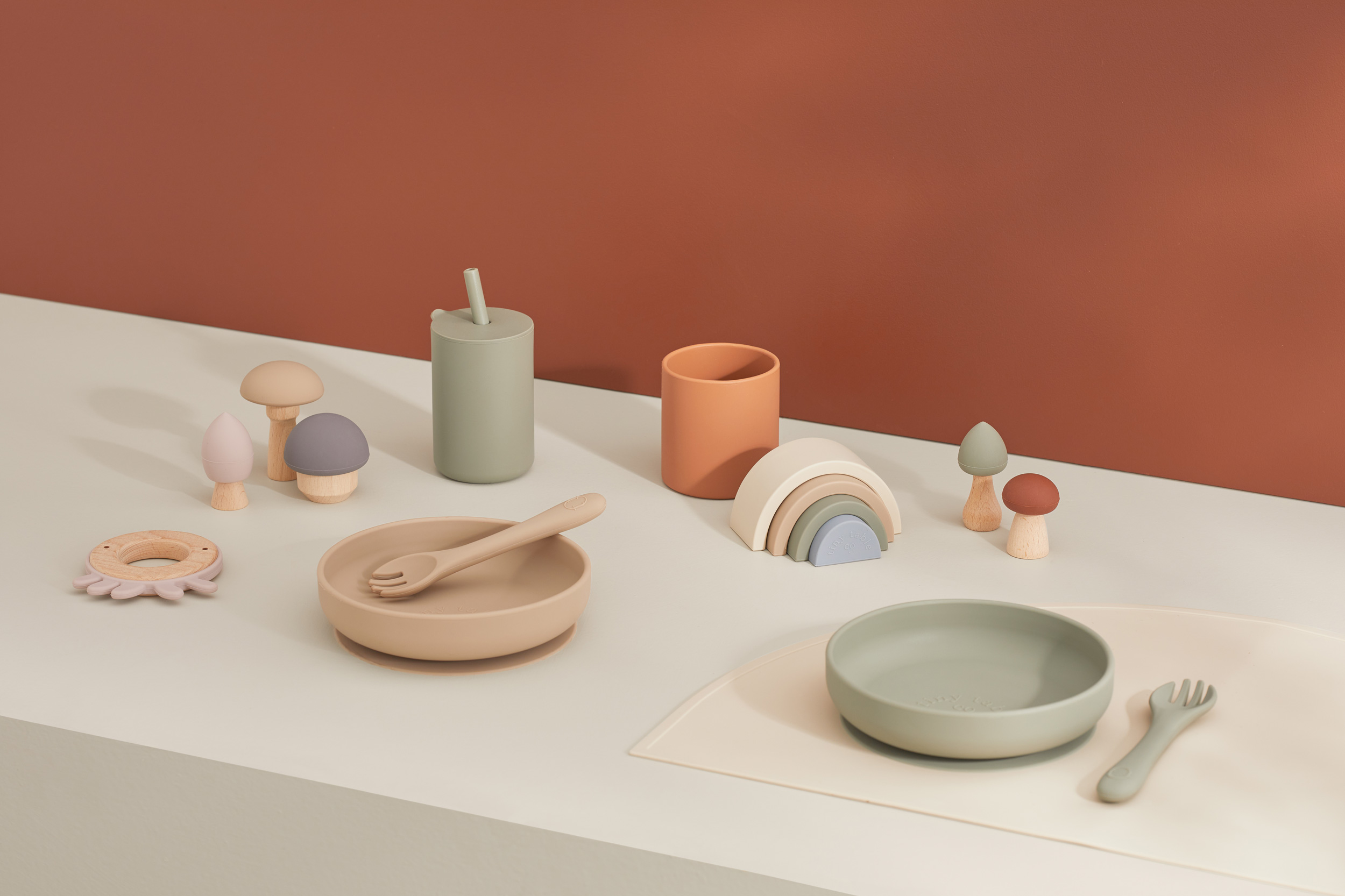Neutral-toned tableware and toys including plates, cups, utensils, mushroom toys, and a wooden rainbow, set on a light surface.