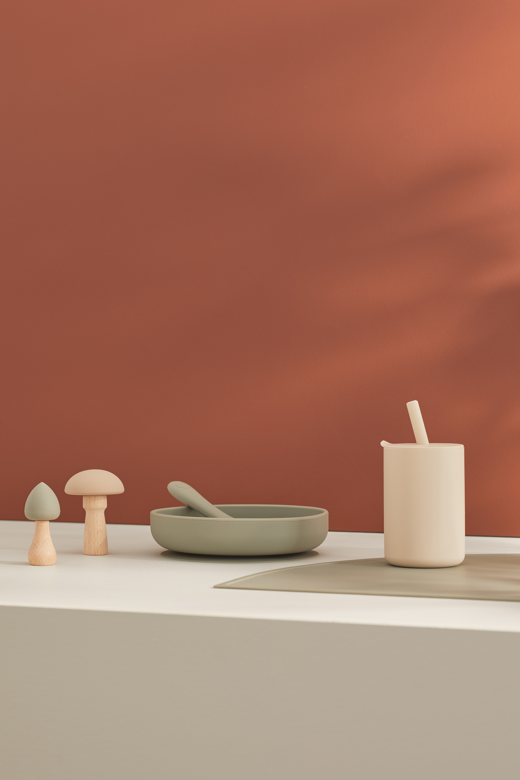 Still life shot of a baby feeding set with a bowl, spoon, sippy cup and two mushroom-shaped toys on a light-colored table.