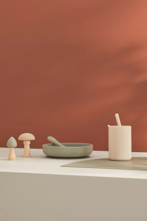 Still life shot of a baby feeding set with a bowl, spoon, sippy cup and two mushroom-shaped toys on a light-colored table.