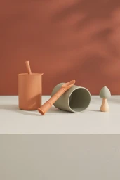 Terracotta and sage green silicone baby cups with a matching spoon and wooden mushroom toy on a neutral surface.