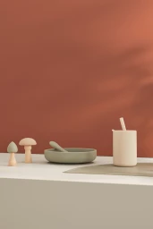 Silicone baby bowl with spoon, straw cup, and wooden toy mushrooms arranged on a ledge against a warm terracotta background.