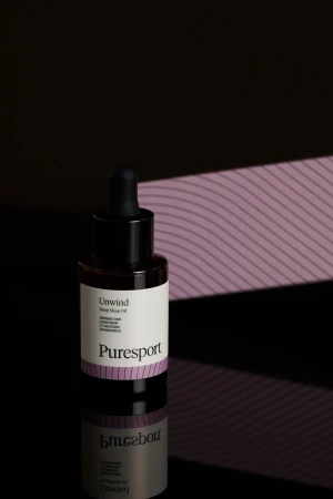Puresport Unwind Sleep Oil product shot. A dark brown glass bottle sits on a reflective black surface against a textured purple backdrop.