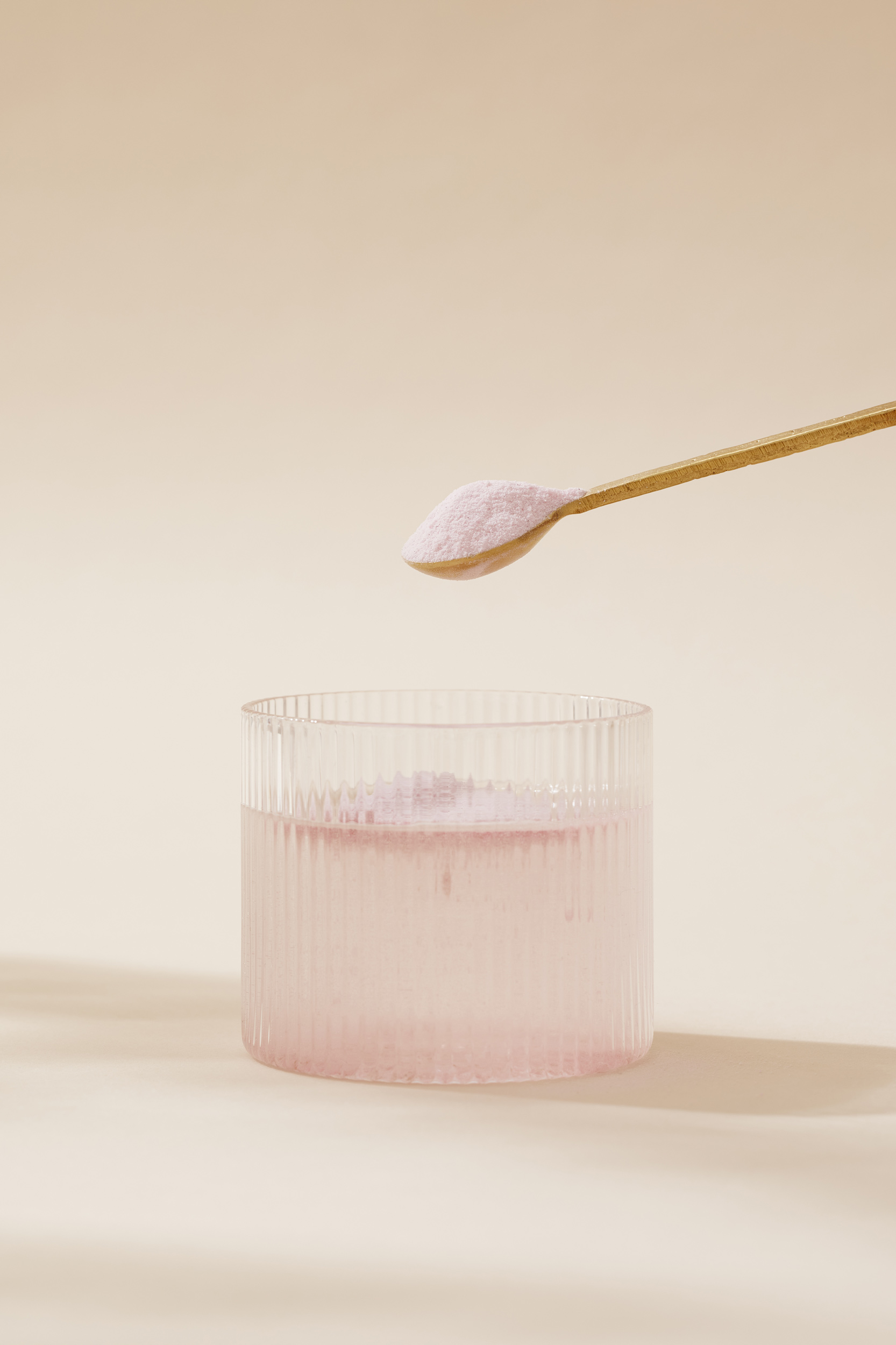 Powder being poured from a wooden spoon into a transparent glass of liquid on a beige background in a minimalist composition.