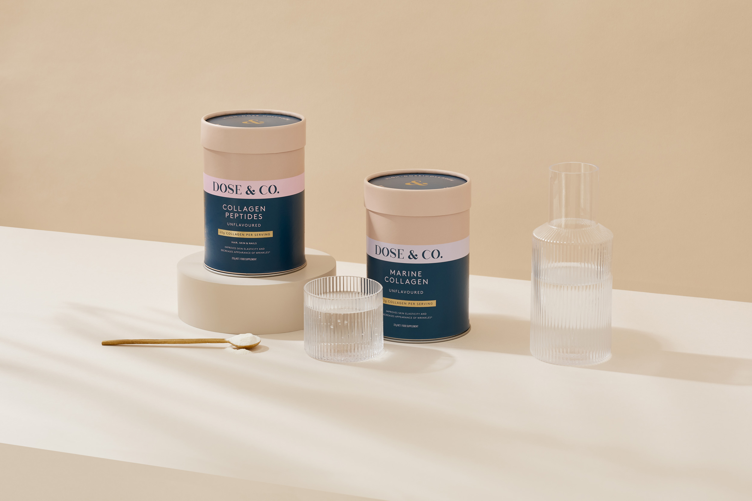 Two product containers and a glass sit on a white surface with a beige background, accompanied by a small spoon and liquid-filled glasses.