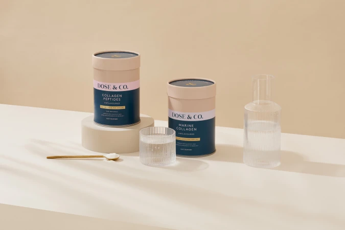 Two product containers and a glass sit on a white surface with a beige background, accompanied by a small spoon and liquid-filled glasses.