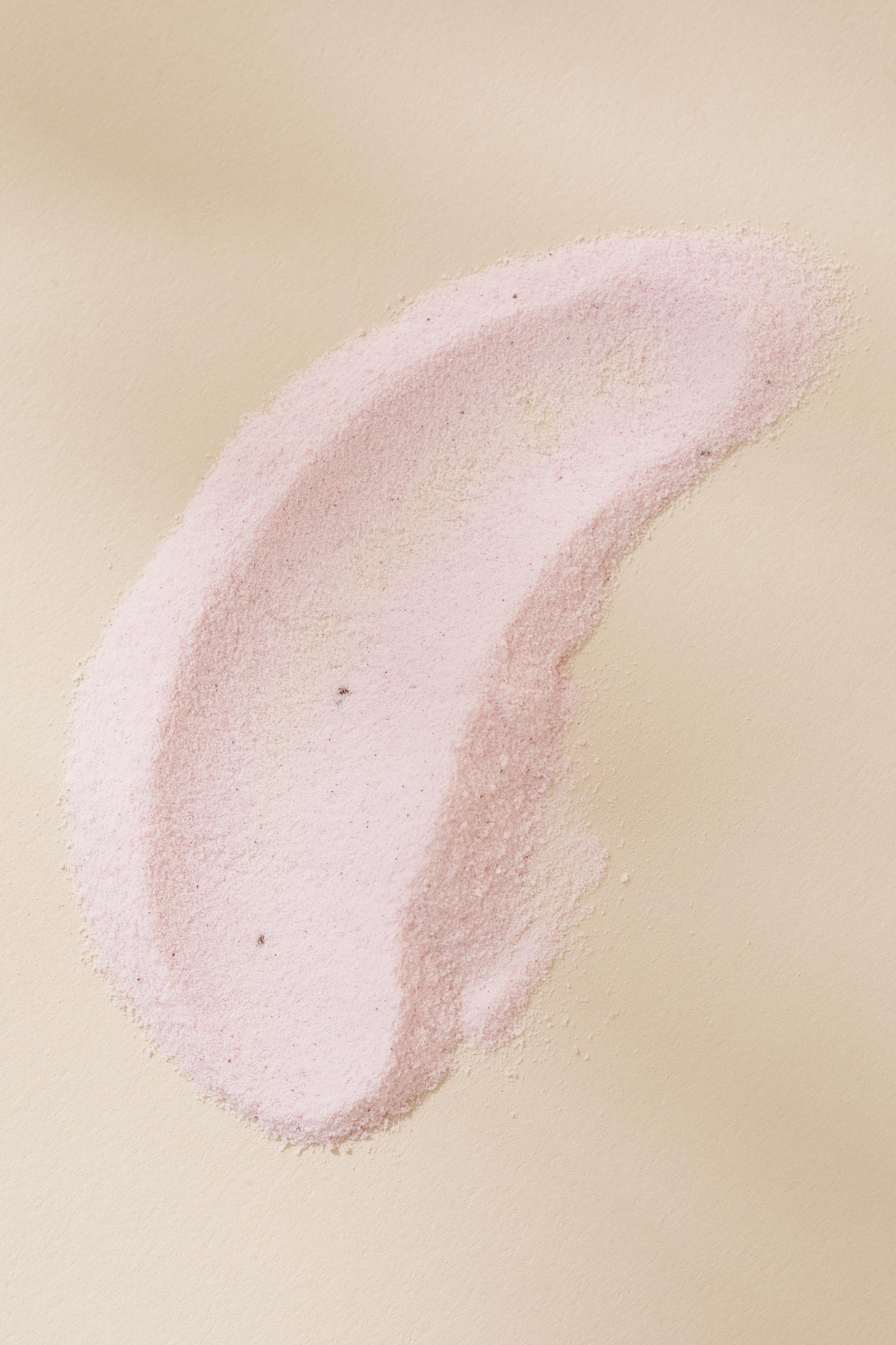 Pale pink cosmetic powder makes an organic, curved shape against a beige background in a studio shot with diffused lighting.