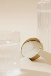 A gold scoop filled with white powder resting on a warm beige studio surface next to a glass of water.