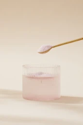 A ribbed glass filled with pale pink liquid beneath a gold spoon holding pink powder against a warm beige background.