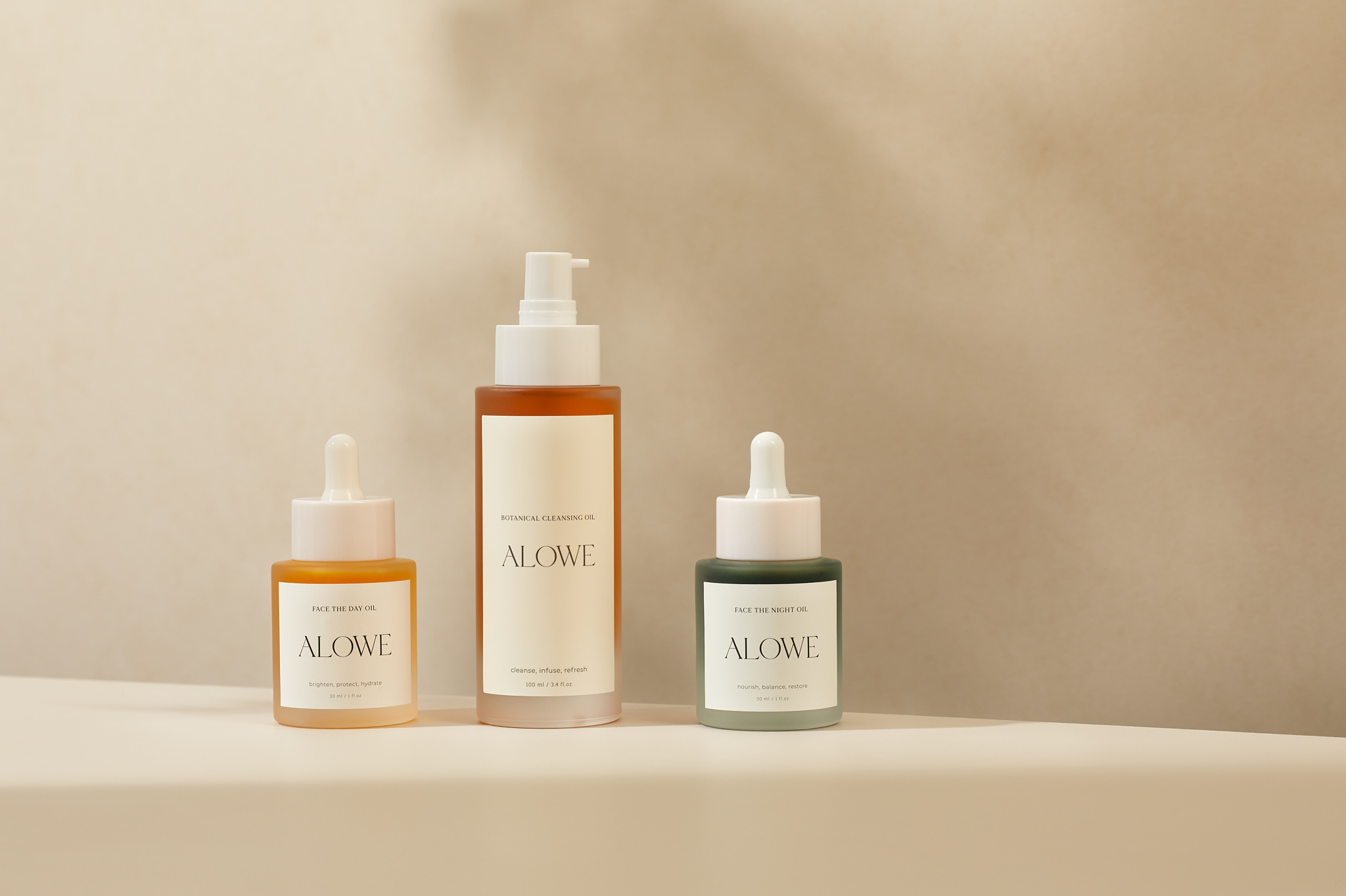 Three Alowe skincare bottles on a white surface against a cream backdrop featuring an oil cleanser and oil treatments. Beauty product shot.