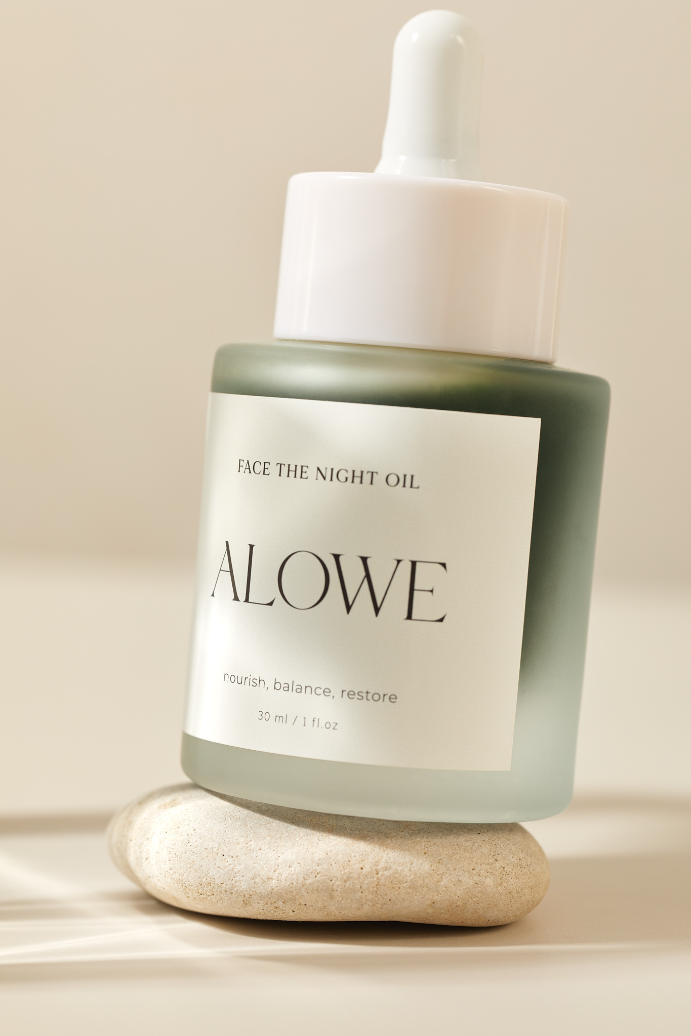 Bottle of Alowe night oil sitting on a light rock. The bottle is a frosted green glass with a white lid and dropper.