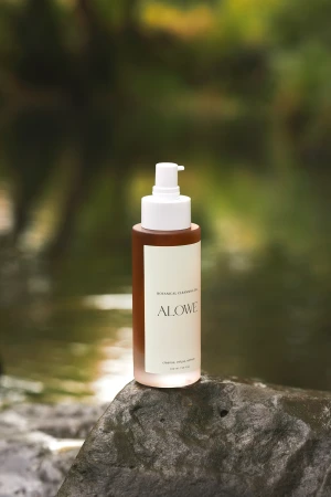 Brown bottle of botanical cleansing oil by ALOWE displayed on a stone, with a green and brown blurred background of trees and foliage.