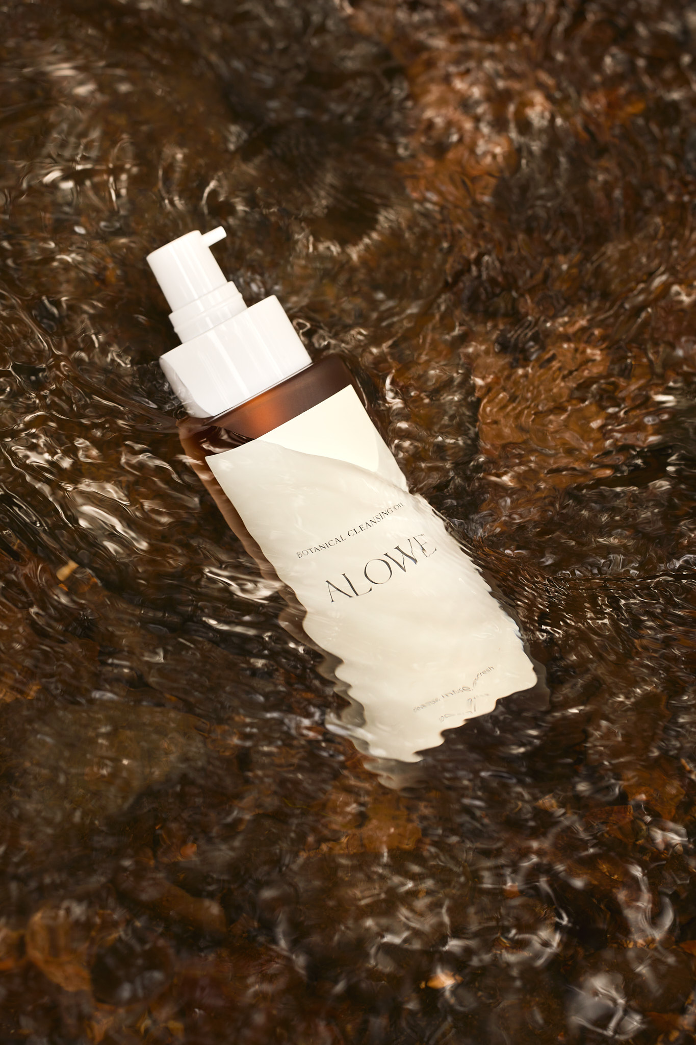 Product shot of a white and brown lotion bottle submerged in shallow water over river rocks, creating ripples and reflections on the label.
