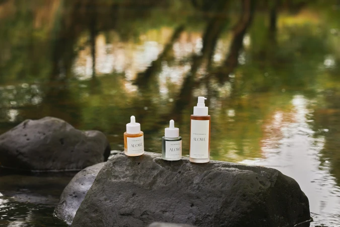 Three Alowe skincare bottles with droppers and a pump dispenser placed on a dark wet boulder in a calm stream.
