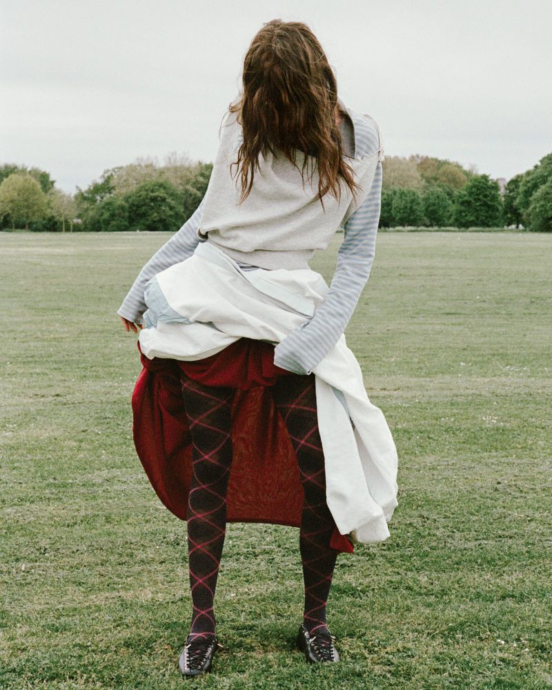 Model in an open field wearing diamond-patterned tights, a layered white and red skirt, and a grey sweater with hair covering the face.