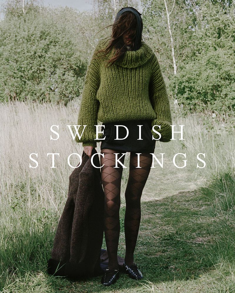 An adult female stands on a grassy path in a green sweater and black stockings, holding a coat in an outdoor setting.