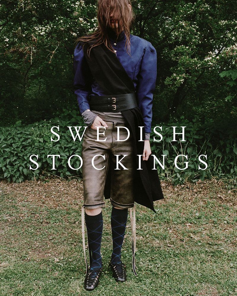 Fashion editorial shot of an adult female model wearing Swedish Stockings brand hosiery, posed in a lush, green outdoor setting.