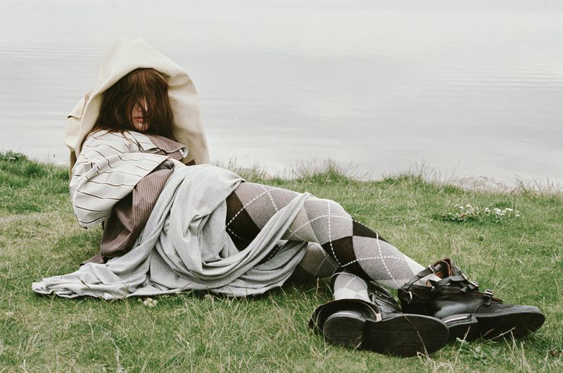 Woman in argyle tights sits on grass in layered clothing, with black leather boots next to a body of water, creating a fashion editorial.