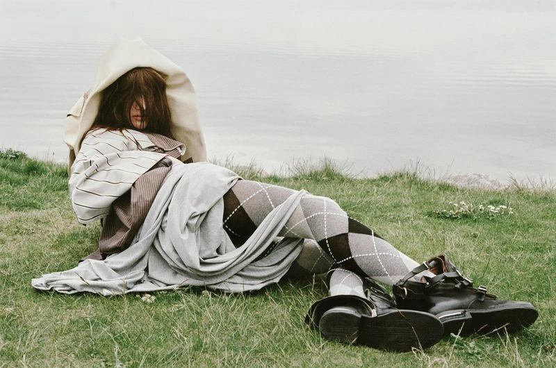 Woman in argyle tights sits on grass in layered clothing, with black leather boots next to a body of water, creating a fashion editorial.