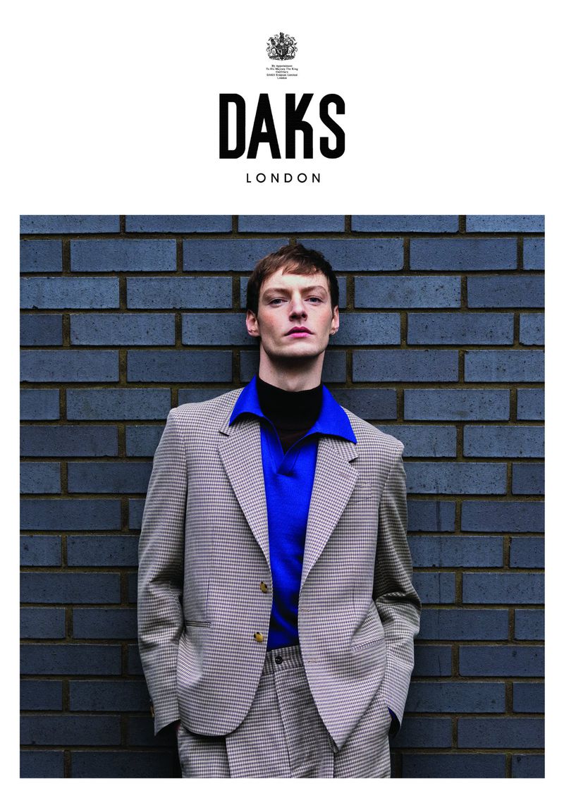 A male fashion model poses against a brick wall in a suit and blue polo shirt for a DAKS London fashion editorial.