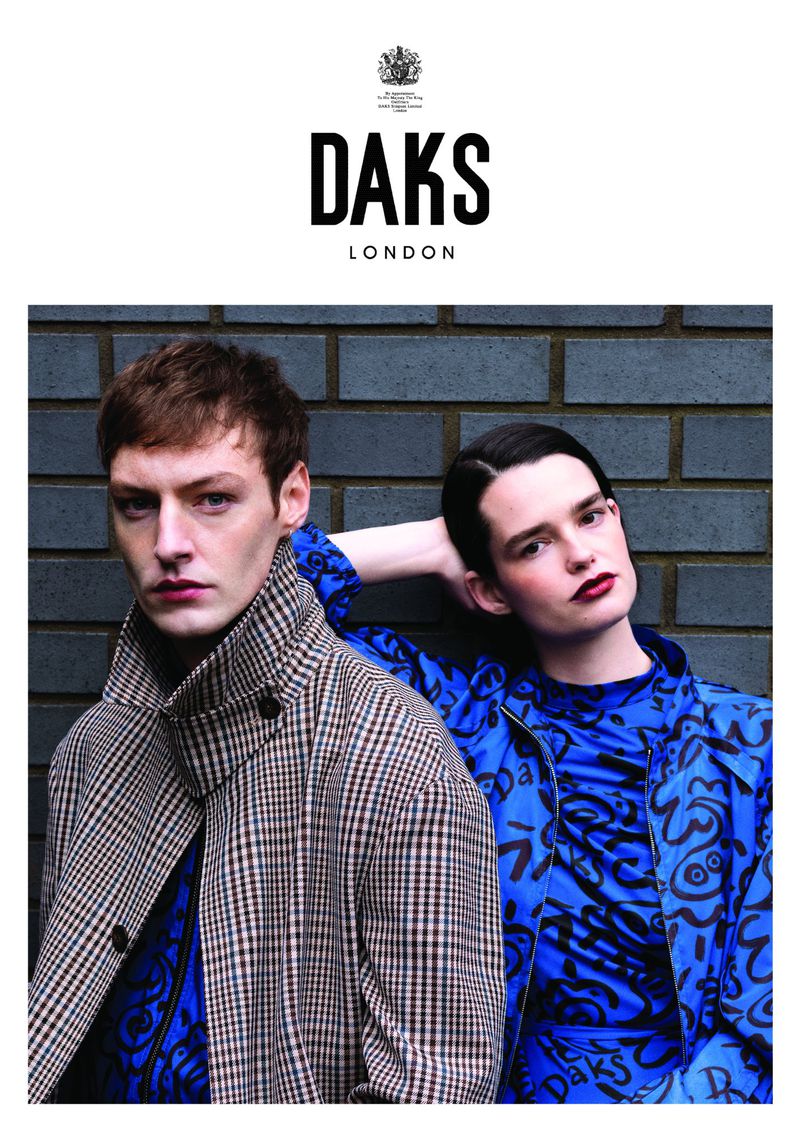Fashion advertisement featuring a young adult male in a plaid coat and a young adult female in a blue printed jacket, posing against a brick wall.