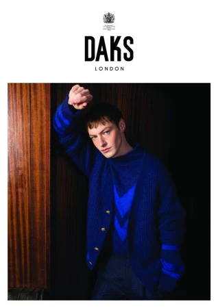 An adult man leans against a dark wooden wall, wearing a blue striped cardigan and sweater, with the DAKS London logo overhead.