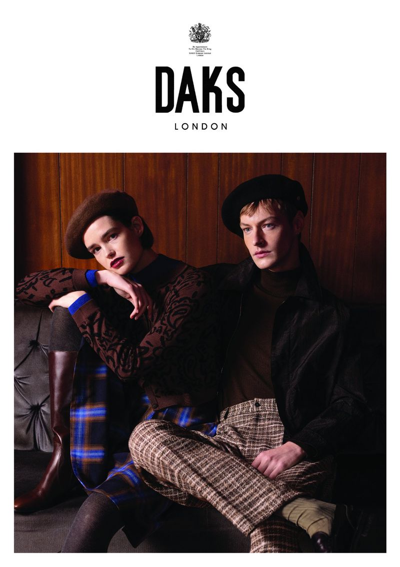 Two models pose for a fashion advertisement wearing berets and patterned clothing, seated on a tufted bench in warm, soft lighting.