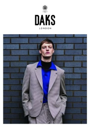 Male model in a patterned grey blazer, blue shirt and black turtleneck leaning against a dark brick wall