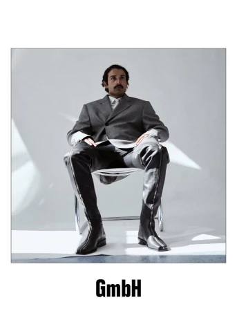 Portrait of a man in a gray suit and black over-the-knee boots sitting on a transparent chair in a photo studio setting.
