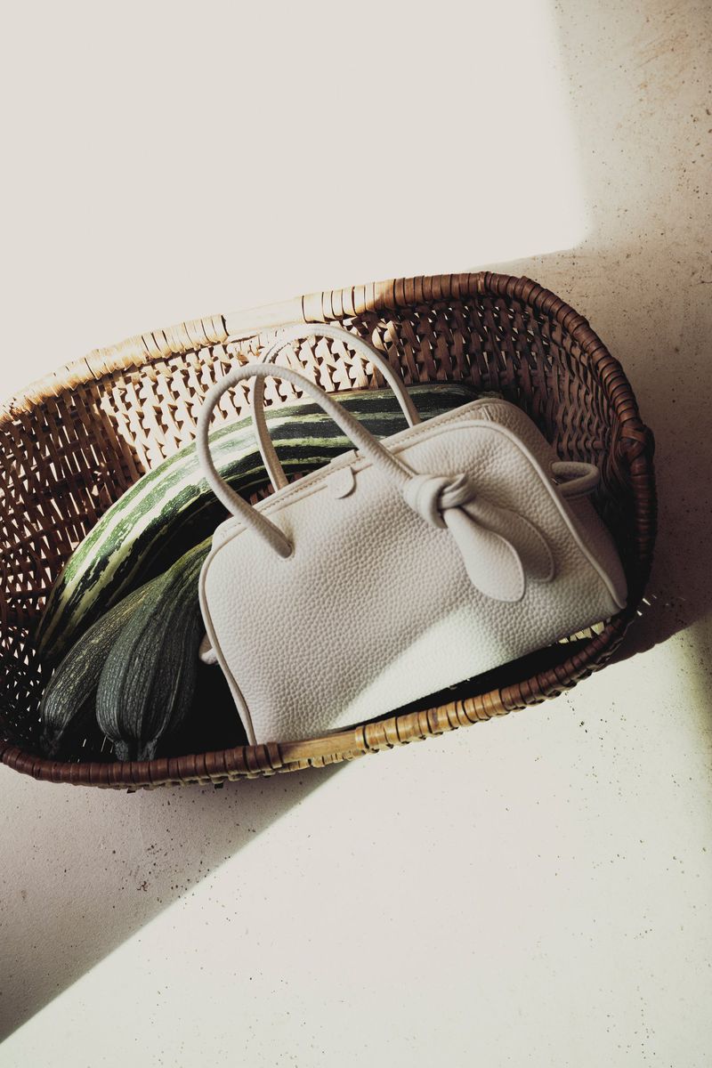 Fashionable handbag in a woven basket, with green squash; studio lighting highlights the bag's texture and shape, creating an appealing aesthetic.