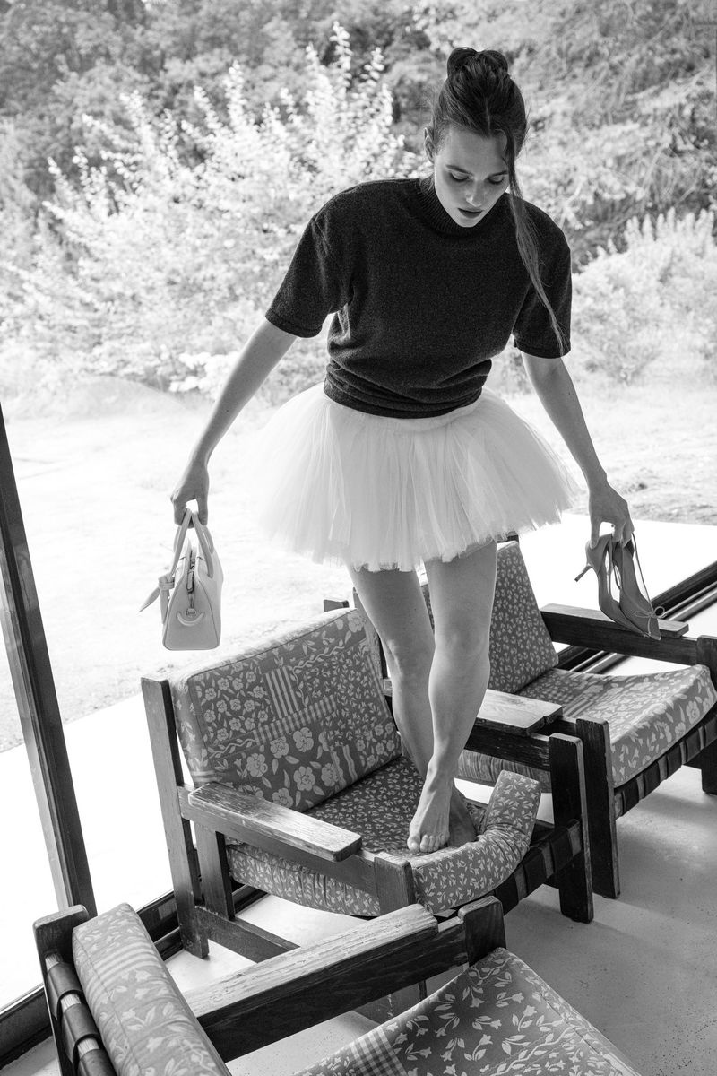 Black and white photo of a woman standing barefoot on armchairs, wearing a tutu and sweater, and holding a purse and high heels.