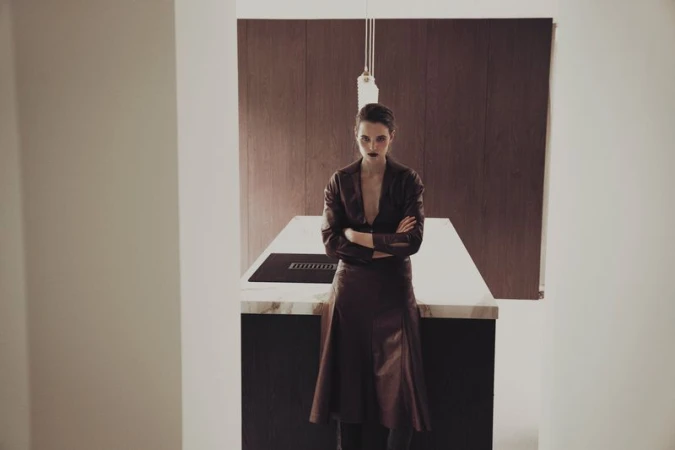 Adult woman in a brown leather suit stands with arms crossed in an indoor studio setting, near a kitchen counter and brown paneling.