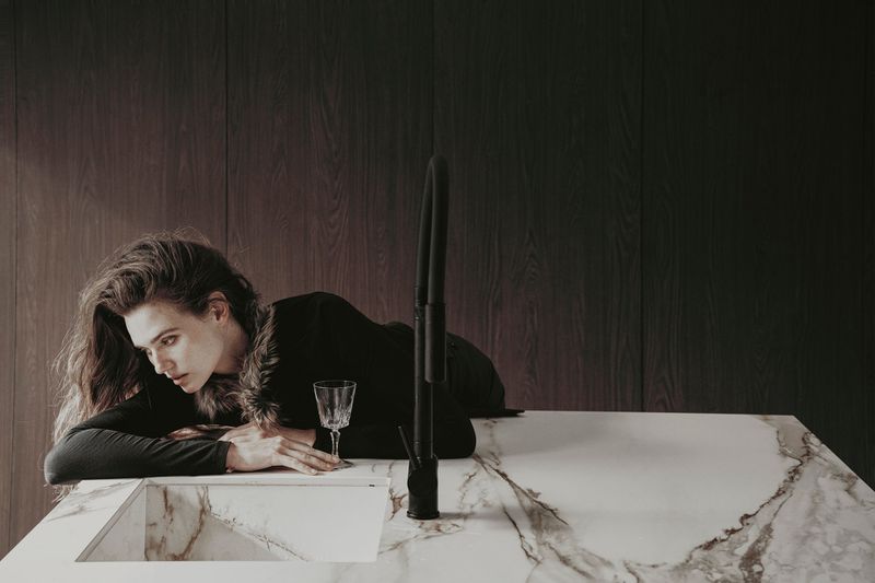 Woman with long hair poses at a counter with a wine glass in a dimly lit interior with a dark wood wall and marble countertop.