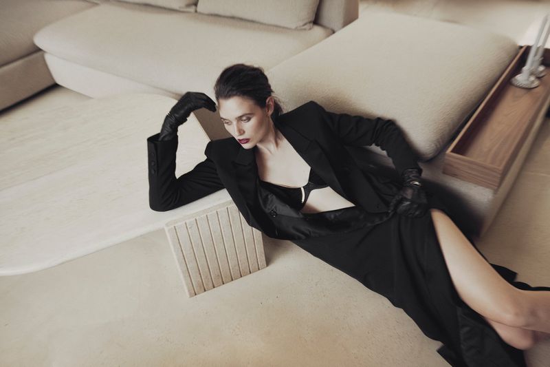 A woman in a black blazer and skirt poses on a white floor near a cream couch, exuding a modern and sophisticated aesthetic in an indoor setting.