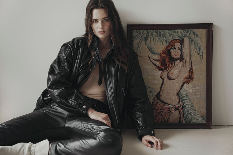 Young woman in a leather jacket and trousers sits next to framed vintage artwork of a pin-up girl, posing in an indoor setting.