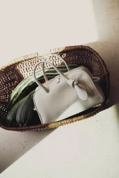 A cream leather handbag resting inside a woven wicker basket next to striped squash under direct sunlight.