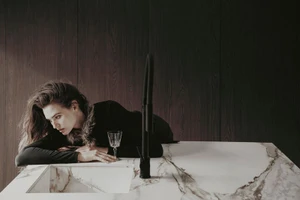 Model lying on a white marble kitchen counter next to a black faucet and crystal glass in a dark wood interior.