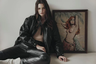 Model seated on the floor wearing a black leather jacket and trousers next to a framed tapestry of a woman
