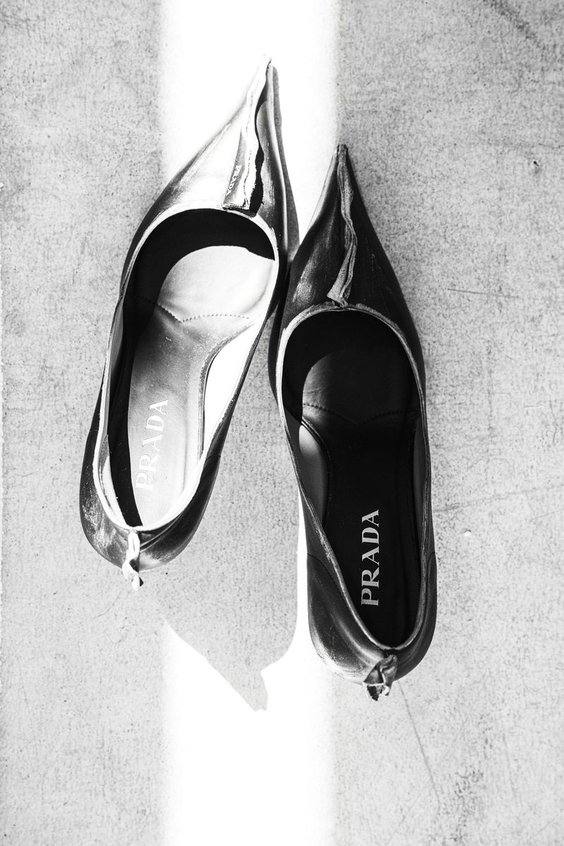 Elevated shot of a pair of Prada high heel shoes, one silver and one black, angled on a bright concrete surface, casting sharp shadows.