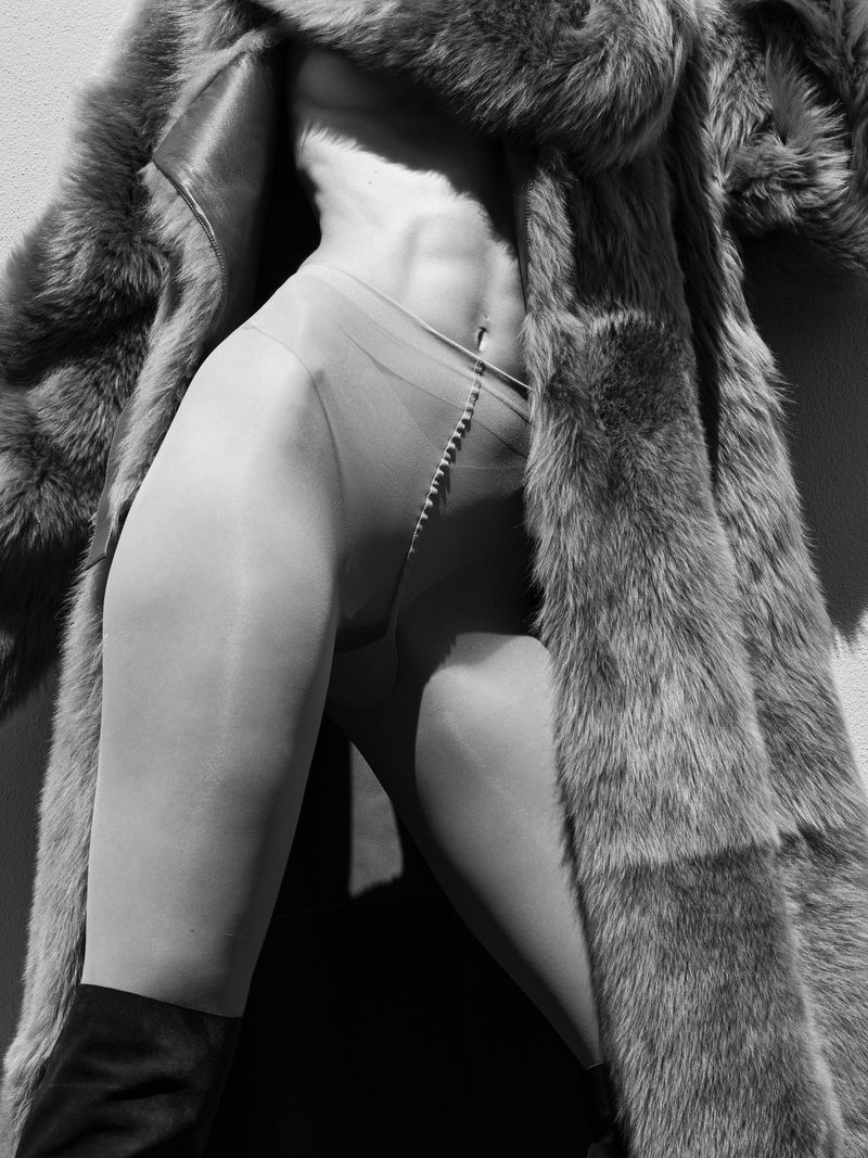 Black and white studio fashion shot of a woman's torso, legs, and shoulders wearing hosiery, fur coat, and boots against a wall.