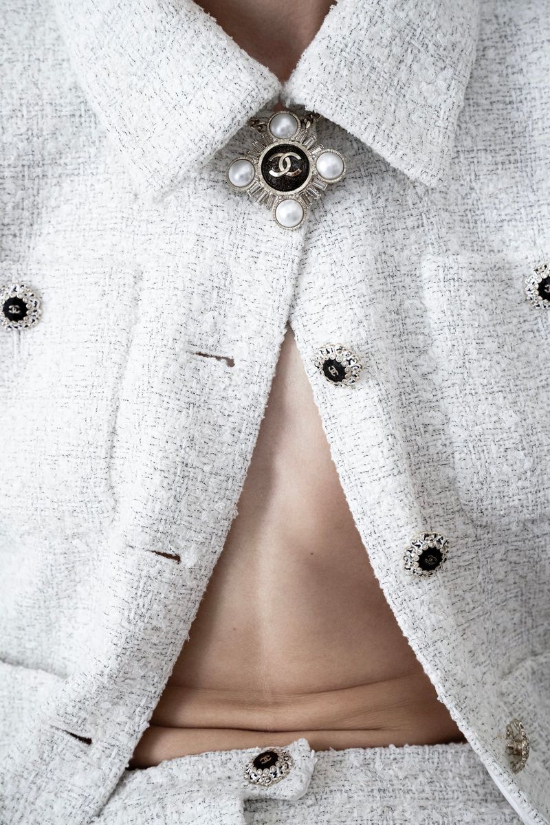 Close-up of a person wearing a white tweed jacket with Chanel logo button and a cross brooch with pearls. Fashion shot.