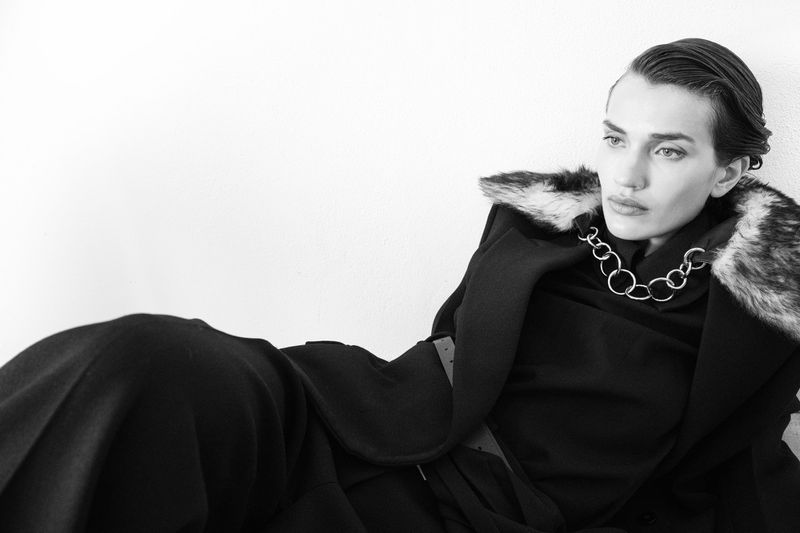 Indoor portrait of an adult woman with wet-look hair in black coat with fur collar and silver necklace, against a white background.